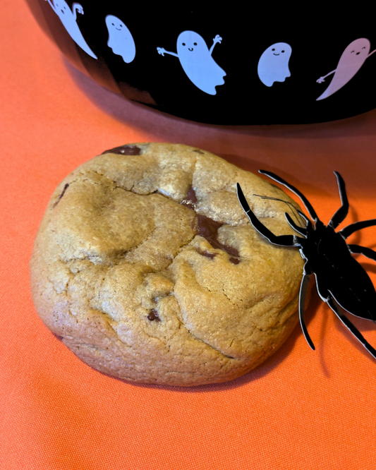 Chocolate chip cookie with a spider on an orange surface, next to a Halloween-themed bowl.